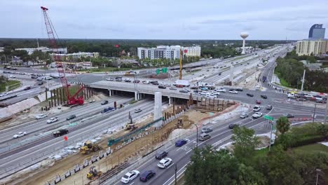 toma aérea de drones con panorámica lenta en ascenso de una concurrida intersección de salida de autopista con paso elevado y zona de construcción de carreteras que muestra tráfico vehicular, equipos de construcción de carreteras y edificios en segundo plano