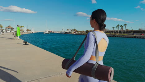 Brunette-woman-walking-on-the-boardwalk-with-yoga-mat-and-sportswear-on-a-sunny-day,-rear-side-view