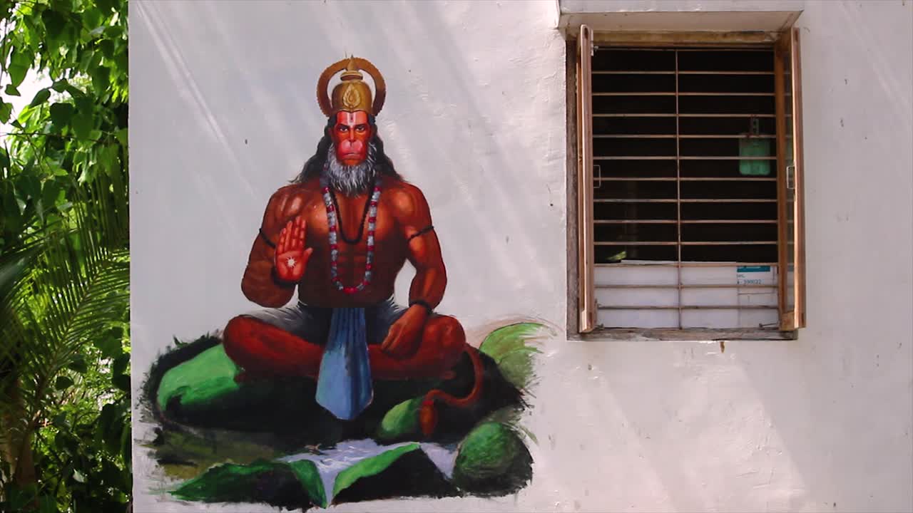 Close-up of Lord Hanuman mural on white house wall near window. Bright Hindu spiritual artwork with natural light and green trees.