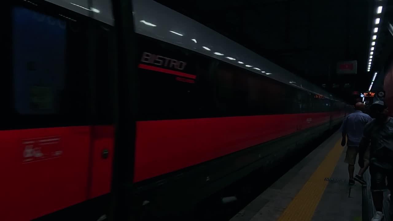People walk beside a stationary Frecciarossa train at a dimly lit platform.