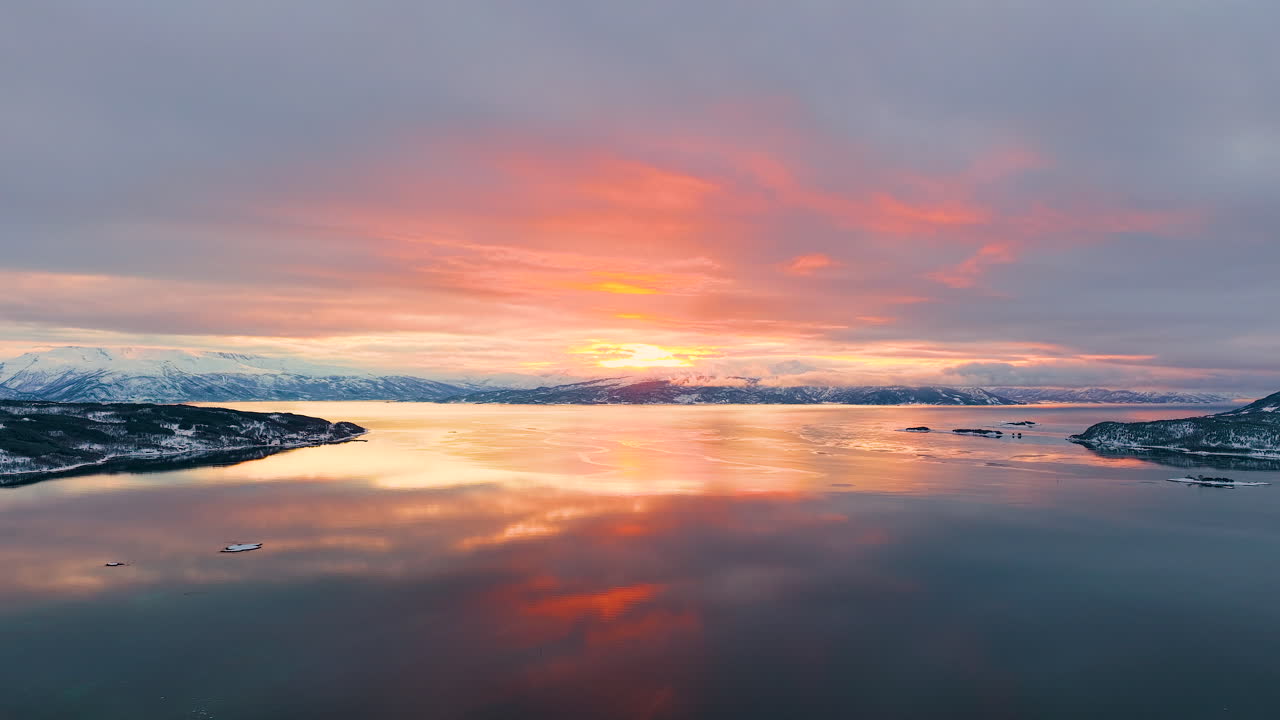 Breathtaking aerial view of a vibrant orange sunset reflecting on the calm waters of a Norwegian fjord, surrounded by snowcovered peaks in the Lofoten archipelago