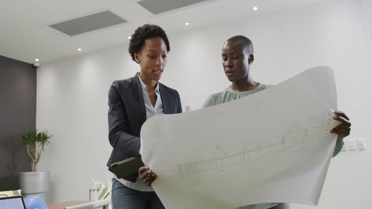 Two african american female architects discussing blueprints in office, in slow motion