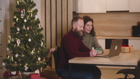 vista lateral de una pareja que compra en línea con una tarjeta de crédito usando una computadora portátil sentada en una mesa cerca de un regalo en una habitación decorada con un árbol de navidad 1