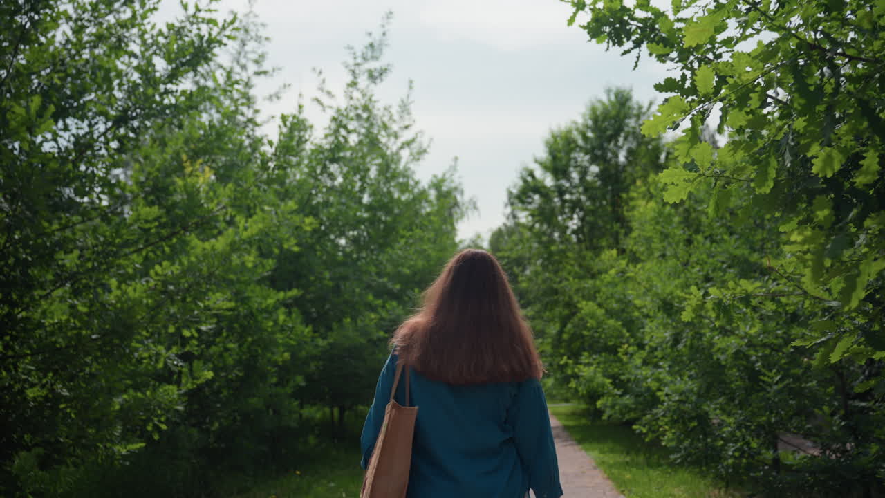 파란색 상의와 흰색 반바지를 입고, 어깨에 자수 가방을 멘 채 햇빛이 비치는 공원 골목을 따라 걷는 학생의 뒷모습, 주변은 무성한 녹색 나무로 둘러싸여 있어요.