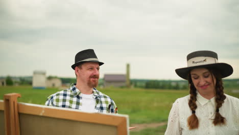 a couple stands in a green field with an art board in front of them. the man is wearing a checked shirt and a hat, while the woman is dressed in a white dress and a wide-brimmed hat