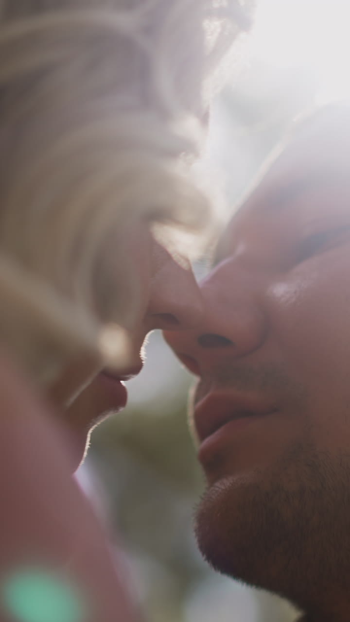 Loving young groom kisses pretty blonde woman bride holding elegant wedding bouquet in green city park closeup low angle shot slow motion
