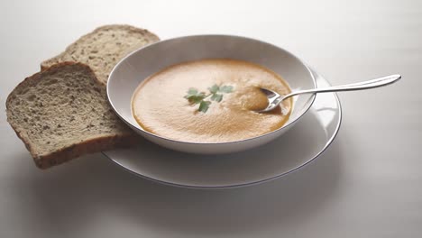 steaming hot butternut squash soup with a sprig of parsley floating on the surface, in a white bowl on a white table with sliced bread and a spoon resting in the bowl.