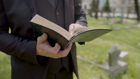 close up view of man hands holding a bible and reading in front of a tombstone in a graveyard