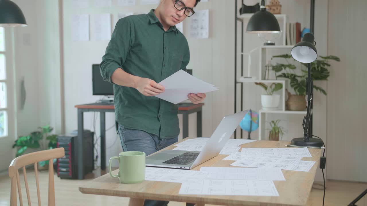 Creative Asian Designer Works On A Storyboard, Looks At His Sketches And Concepts, Choosing The Best Drawings For His Project. Video Editing, Comics Compilation, Application Ui Or Game Plot