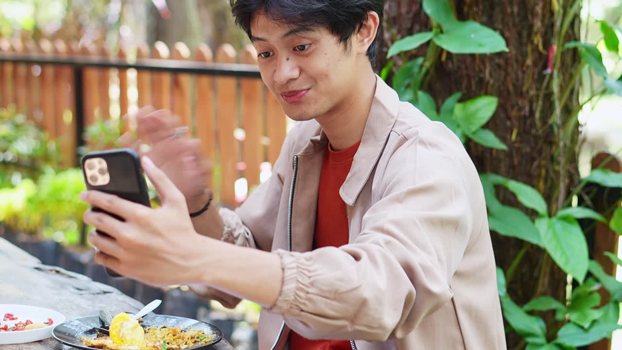 Happy young asian man sharing fun moments online with phone selfie and thumbs up enjoying outdoor cafe life