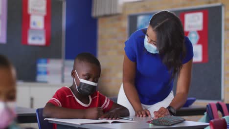 Diverse-female-teacher-helping-schoolboy-with-lesson,-all-wearing-face-masks