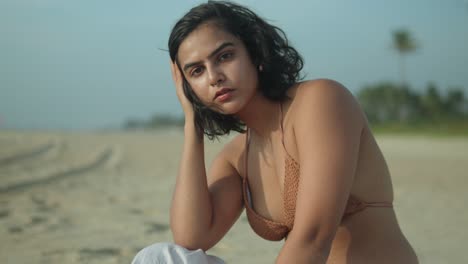 mujer joven posando pensativa en una playa soleada, mano en el cabello, cielo despejado