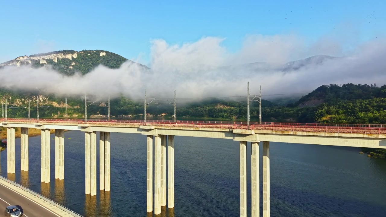穏やかな貯水池を横断する高架高速道路の橋にかかる日の出。森林に覆われた丘陵には霧が漂い、連続するコンクリート橋脚、暖かい光、穏やかな水景、そして遠くの地平線が広がる。