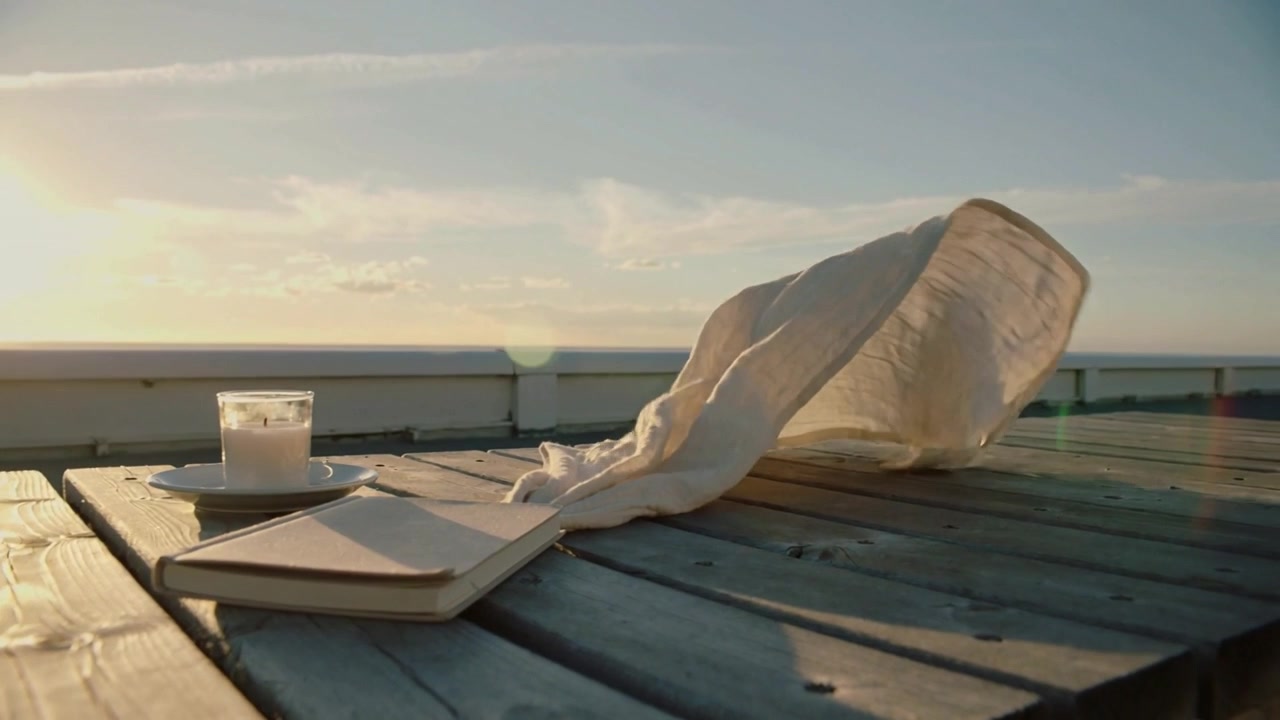 Breeze picking up, cream cloth billowing across wooden pier at golden hour with notebook, candle