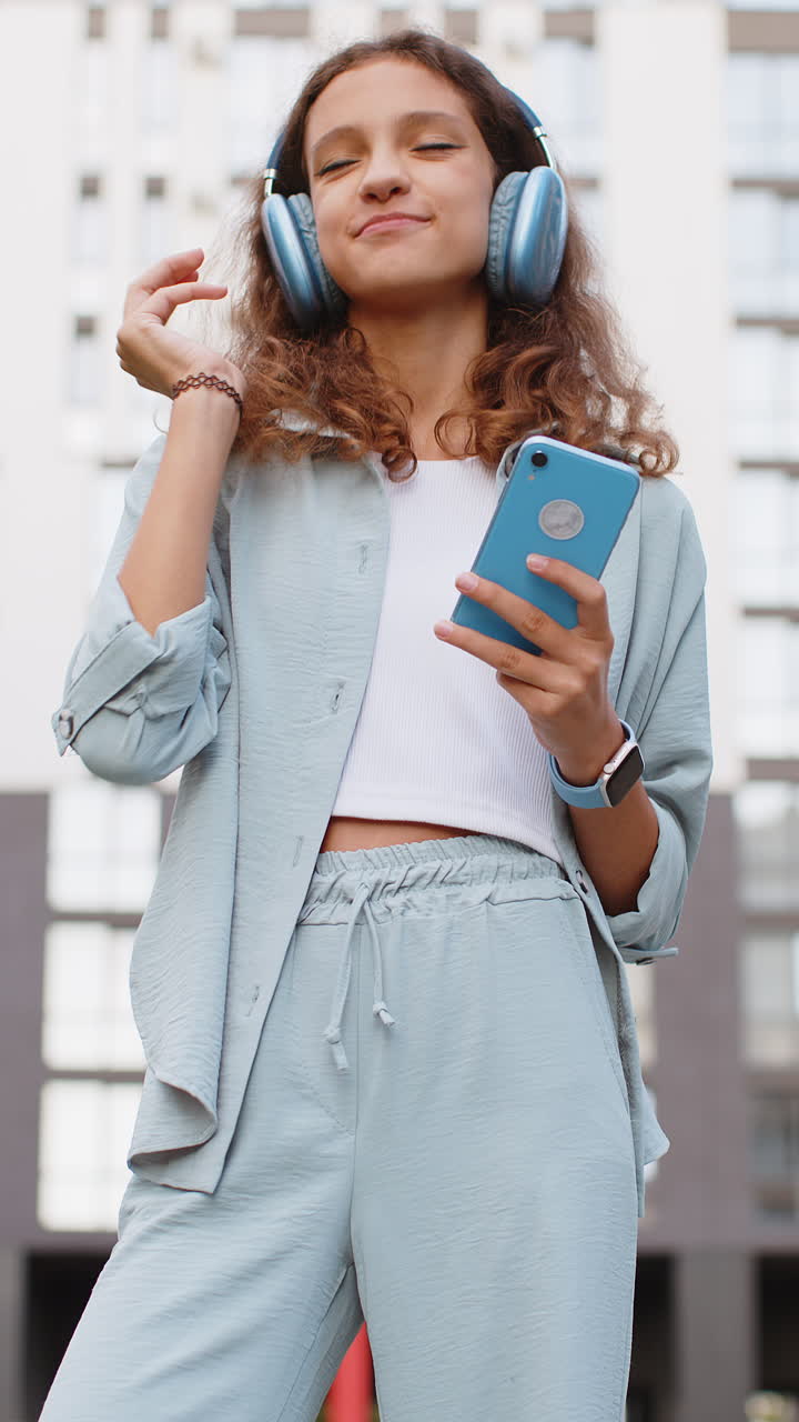 Happy preteen child girl in wireless headphones choosing listen music dancing outdoors city street