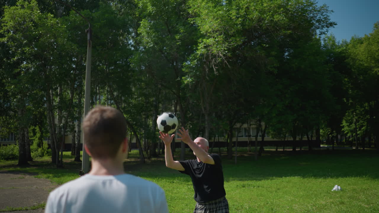 Close-up of an elderly man in a black shirt tossing a soccer ball to his grandson, who heads it back to him, the background features lush green trees and a building