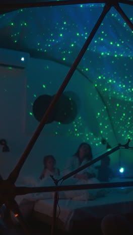 children entertain with mother in bathrobe in dimly lit room. transparent glamping house with starry night sky on ceiling illuminated by contemporary projector
