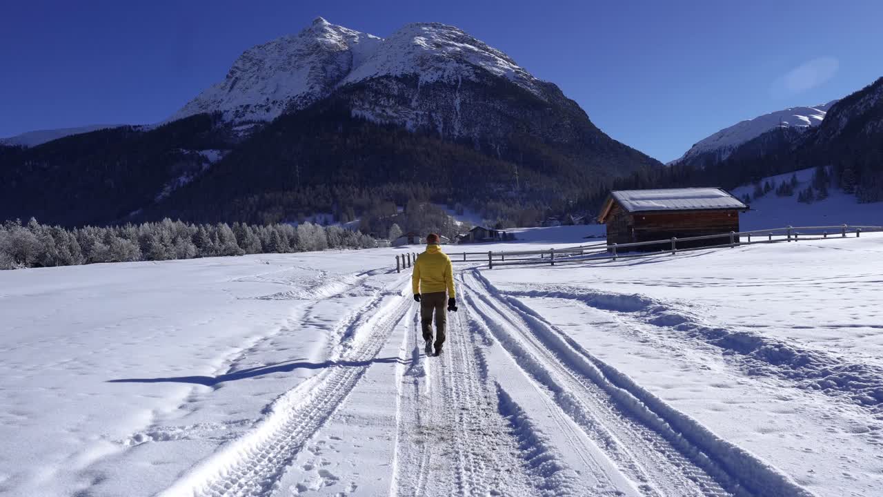 Giovane uomo caucasico con una giacca gialla e un berretto di lana che cammina attraverso un paesaggio invernale innevato con una baita in legno.