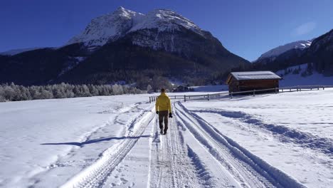 joven caucásico con una chaqueta amarilla y un gorro caminando por un paisaje invernal cubierto de nieve con una cabaña de madera