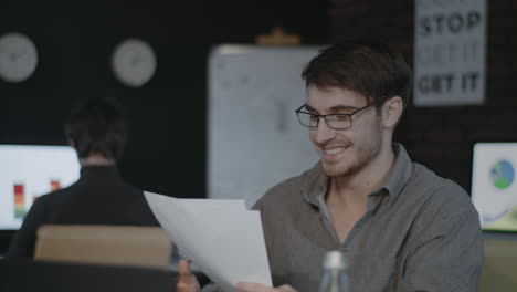 Smiling-business-man-looking-document-paper-on-workplace-in-dark-office