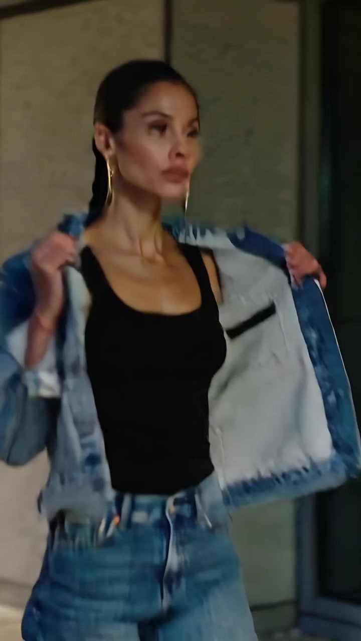Video verticale: La telecamera riprende una modella che cammina in una hall piastrellata di notte, mentre si sistema la giacca di jeans per un servizio fotografico.