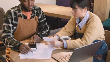 Two-Multiethnic-Coffee-Shop-Owners-Sitting-At-Table-And-Doing-Accounting