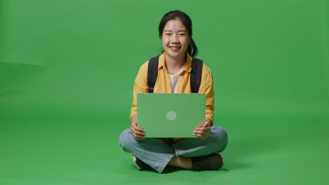 full body of asian woman student with a backpack typing on a laptop and smiling to camera while sitting in the green screen background studio