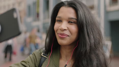 portrait of cheerful young mixed race woman on vacation wearing earphones listening to music taking selfie photo using smartphone