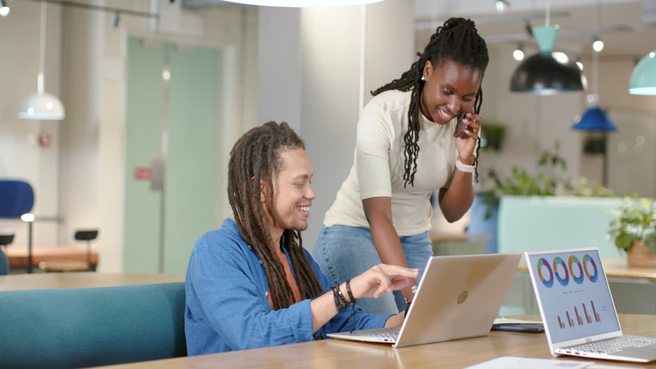 African American team answering phone at table leaning over laptop reviewing charts on right laptop