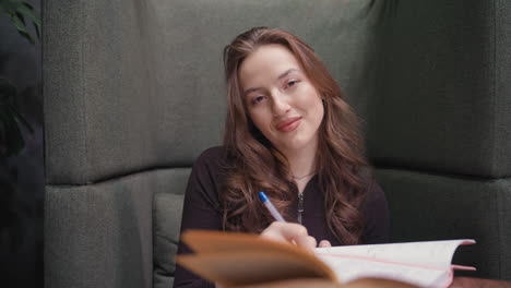 lady with long wavy hair holds pen above open notebook while making soft eye contact with camera, seated in cushioned booth surrounded by calm environment, writing with composed facial expression