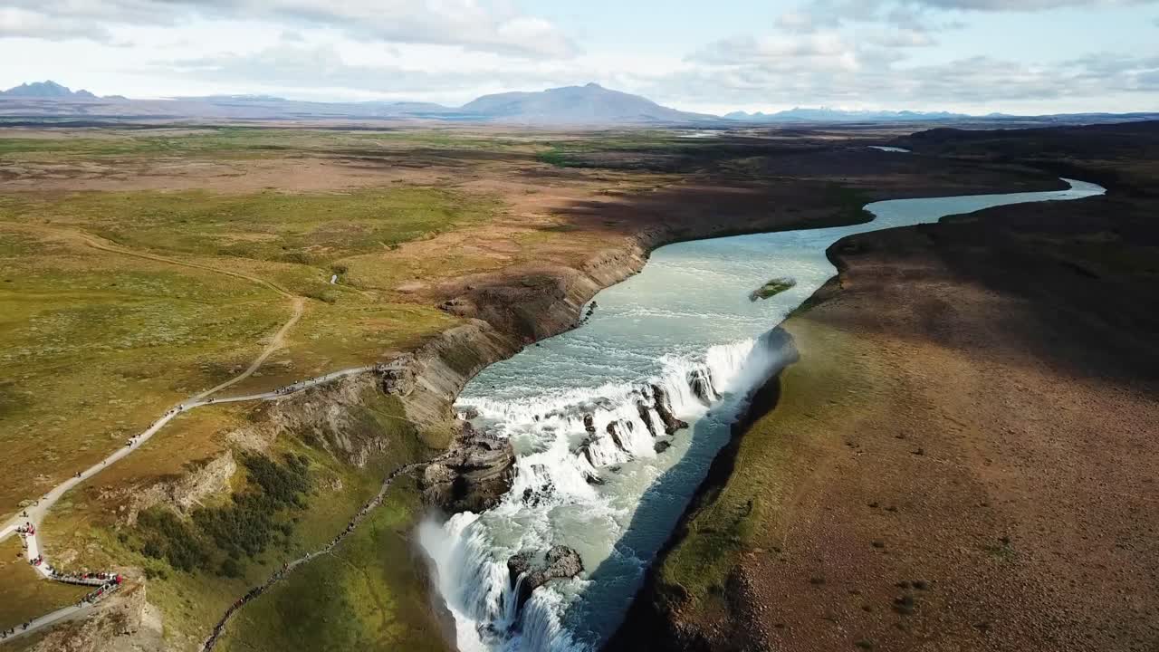 Gullfoss waterfall is cascading down in Iceland, creating a rainbow, in an aerial view, with tourists walking on the trails next to it, and a vast landscape in the background, drone establishing shot