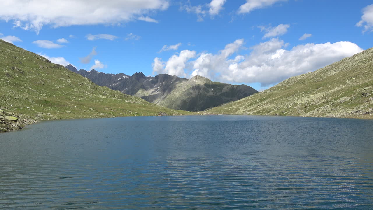Vid udsigt over Merjelensøen med blide krusninger på klart vand, omgivet af grønne alpeskråninger og barske tinder under blå himmel, Valais, Schweiz