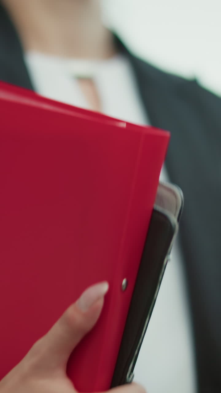 Close up of banker in black suit holding file folder and coffee cup while walking along path with blurred background of parked cars and urban buildings during bright morning commute to office