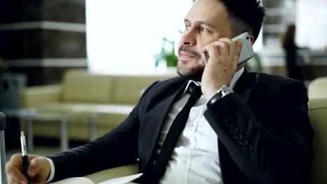 primer plano de un hombre de negocios confiado hablando por teléfono móvil y escribiendo notas en el bloc de notas mientras está sentado en un sillón en la recepción del hotel. concepto de viajes, negocios y personas