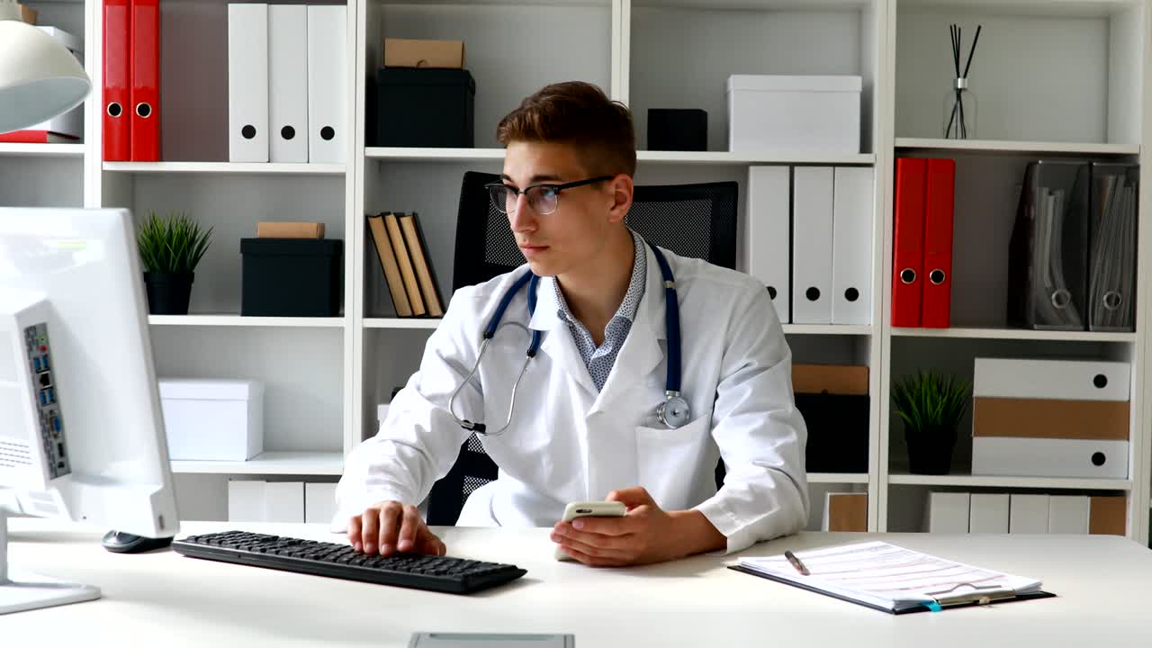 Young doctor holding phone in hand and working at computer