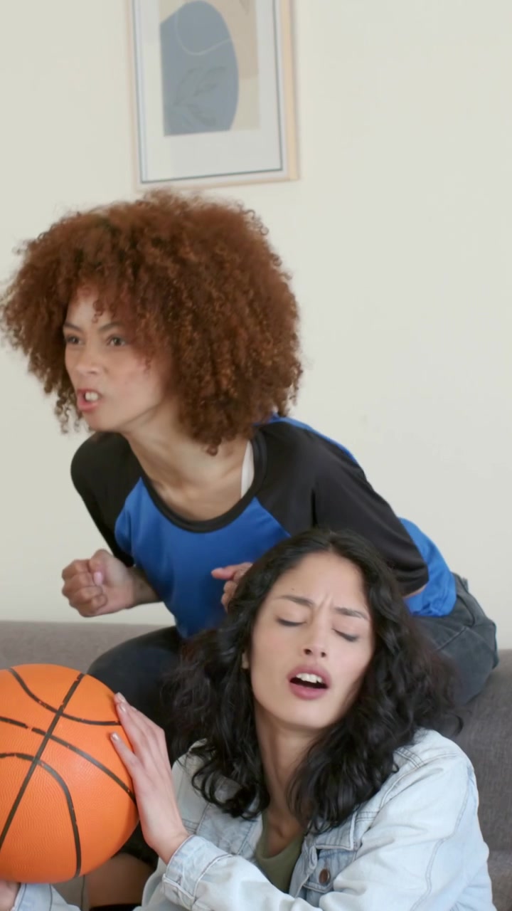 Vertical video: Women in jersey and denim reacting at home watching tense TV, holding basketball