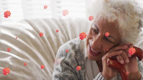 Animation-of-red-roses-over-smiling-biracial-senior-woman-sitting-leaning-on-walking-stick,-at-home