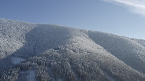 Toma-Aérea-De-árboles-En-Una-Montaña-Cubierta-De-Nieve-Durante-El-Invierno,-Cielo-Azul-Con-Nubes