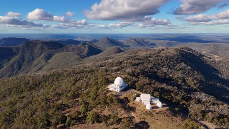 drone glides over a forested mountain ridge, revealing a white observatory dome under bright daylight with scattered clouds and expansive views