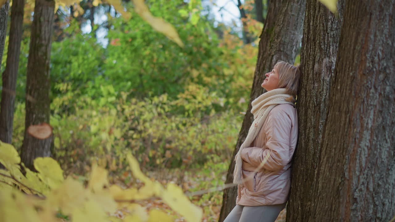 woman in soft beige jacket and scarf leans back against tree trunk in colorful forest, gently lifting hand to catch falling autumn leaf while gazing upward with thoughtful and calm expression