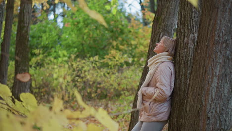 woman in soft beige jacket and scarf leans back against tree trunk in colorful forest, gently lifting hand to catch falling autumn leaf while gazing upward with thoughtful and calm expression