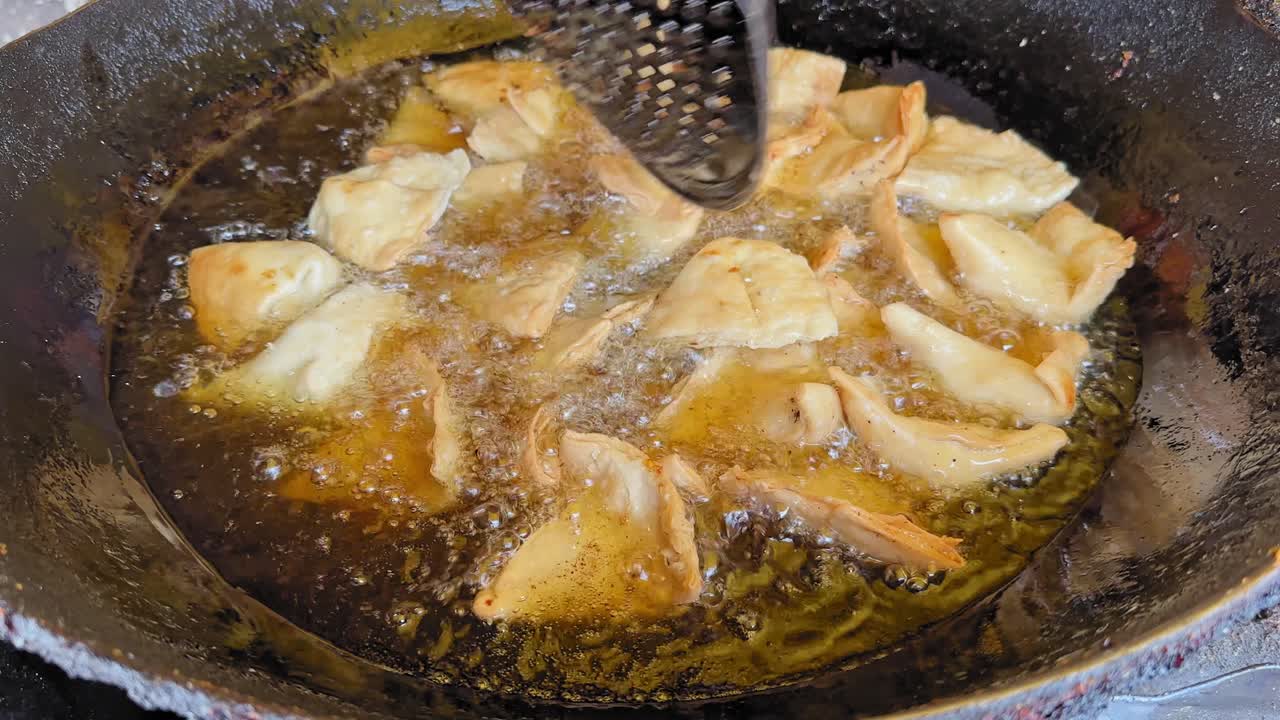 Primo piano di samosa messe in olio bollente all'interno di un wok scuro, superficie gorgogliante e bordi dorati della pasta che si spostano mentre la schiumarola di metallo si muove nell'olio sfrigolante
