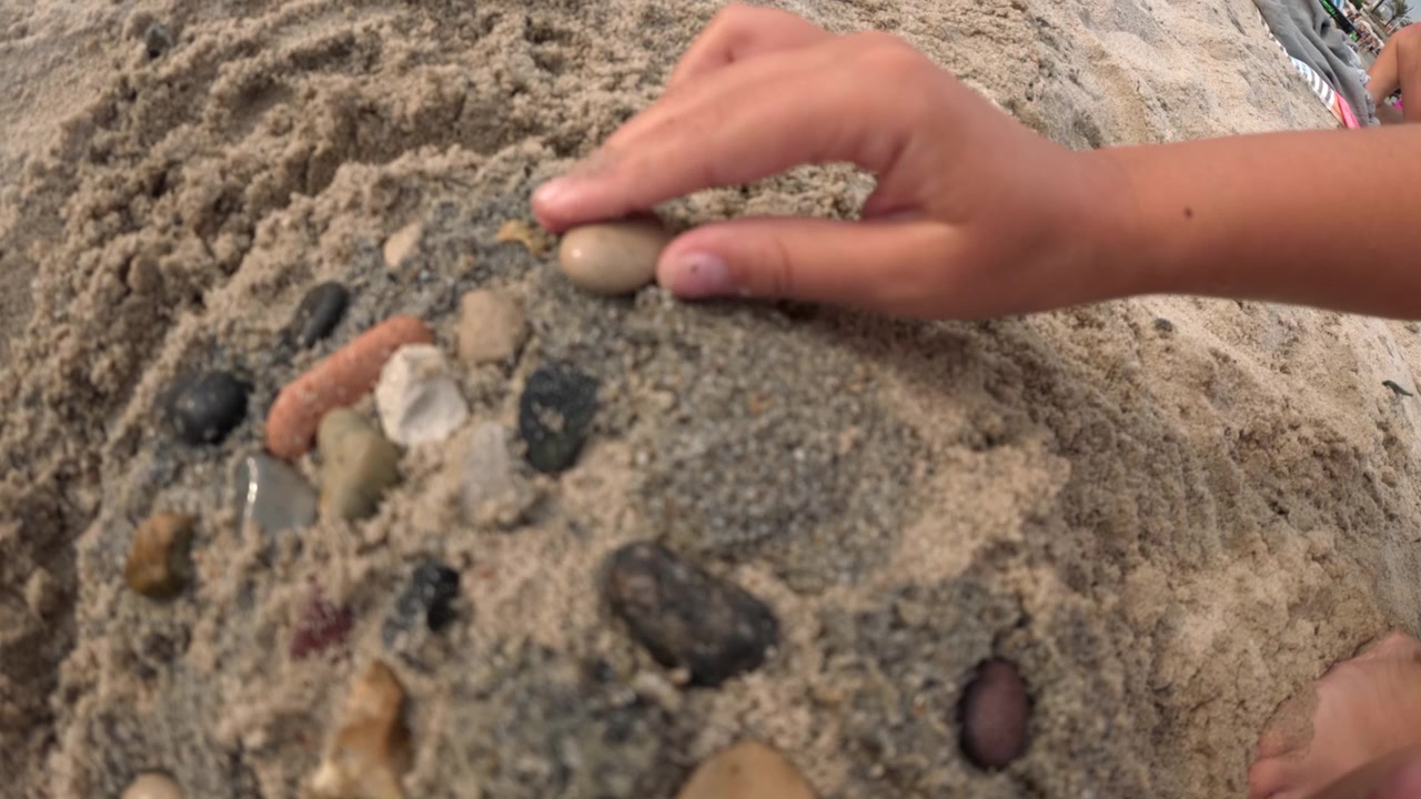 Close-up of a child's hands arranging small colorful pebbles and shells on a sand mound, Golfe-Juan, France