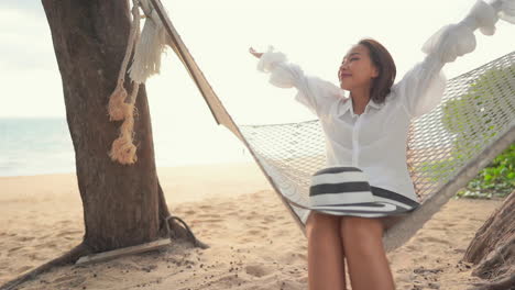 asian woman sitting and swinging in a hammock on a tropical beach in thailand, she is spreading and raising her hands in the air