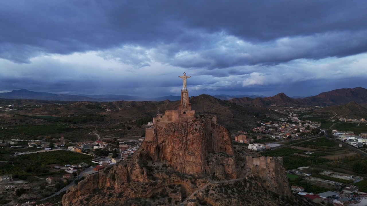 Panorama aereo di un crocifisso monumentale e un'antica fortezza su una collina rocciosa che domina la campagna di Murcia