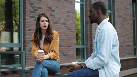 estudiantes caucásicos y afroamericanos hablando y bebiendo café sentados en la calle cerca de la universidad