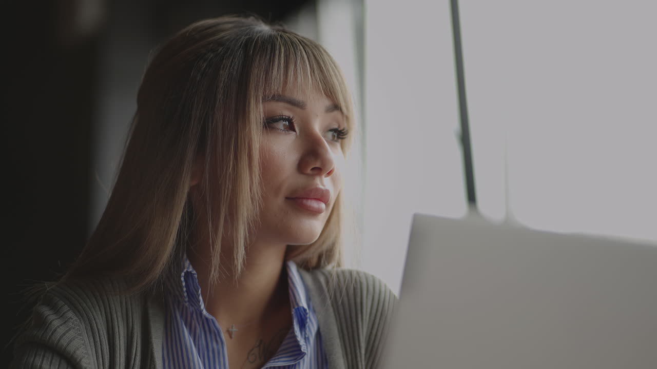 una hermosa joven asiática se le ocurre y resuelve el problema mirando por la ventana. sentada en la computadora portátil para pensar y planear