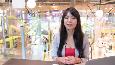 beautiful asian announcer, broadcaster woman reporting news in front of camera with laptop and smartphone on the table in workplace public room background