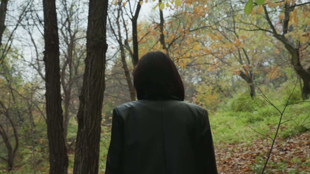 back view woman in leather jacket facing trees contemplative still frame of lone figure at forest edge, soft autumn foliage, quiet path, reflective posture suggesting introspection and cinematic
