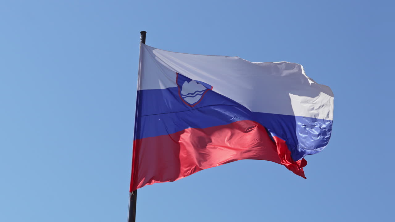 Berrak mavi bir gökyüzüne karşı bir bayrak direğinde dalgalanan Slovenya bayrağı.